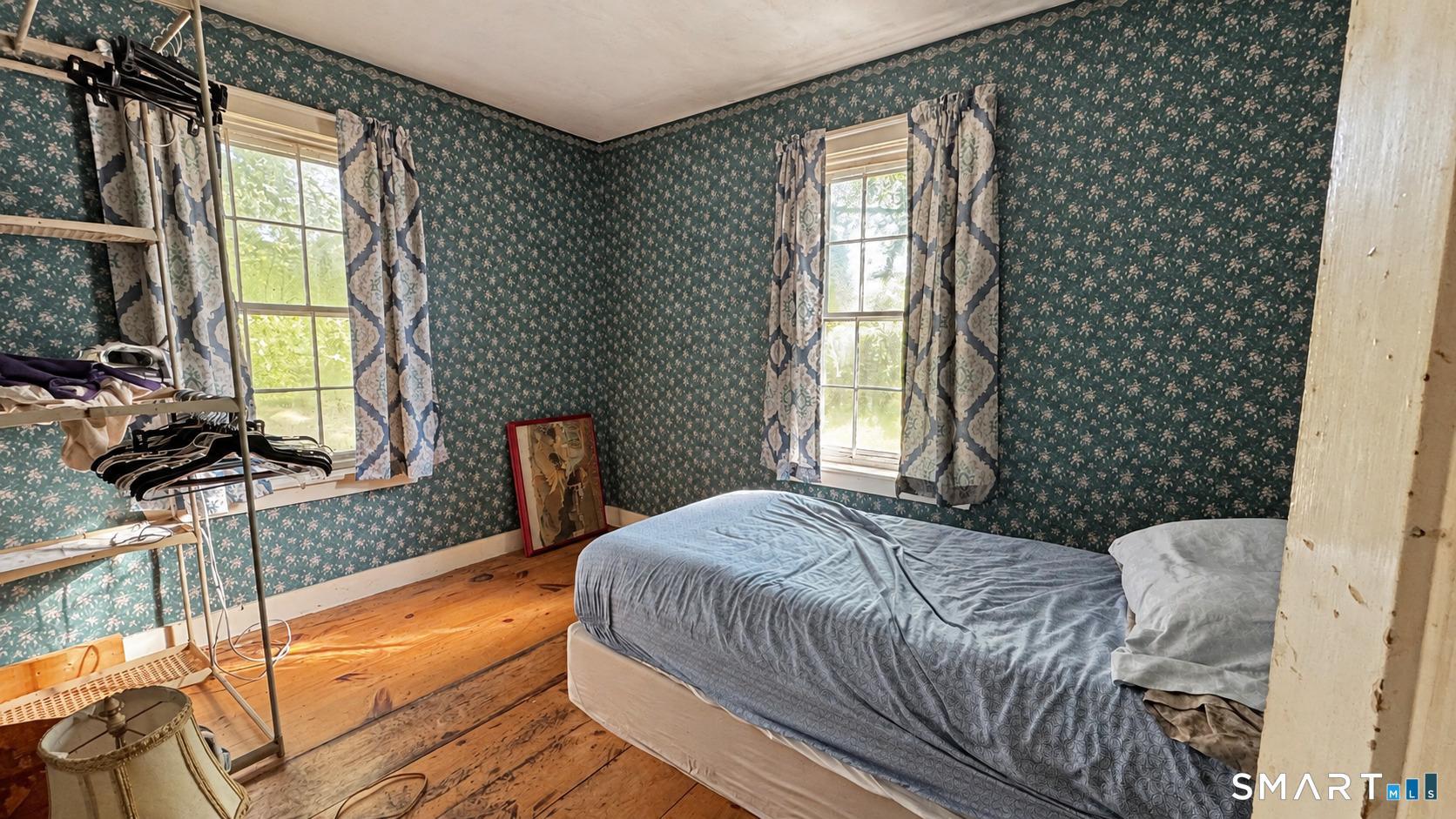 Bedroom with teal floral wallpaper, two curtained windows, and worn wood floors.