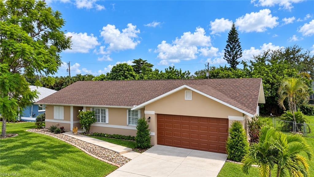 Single-story tan house with a brown roof, two-car garage, landscaped lawn, and tropical trees.