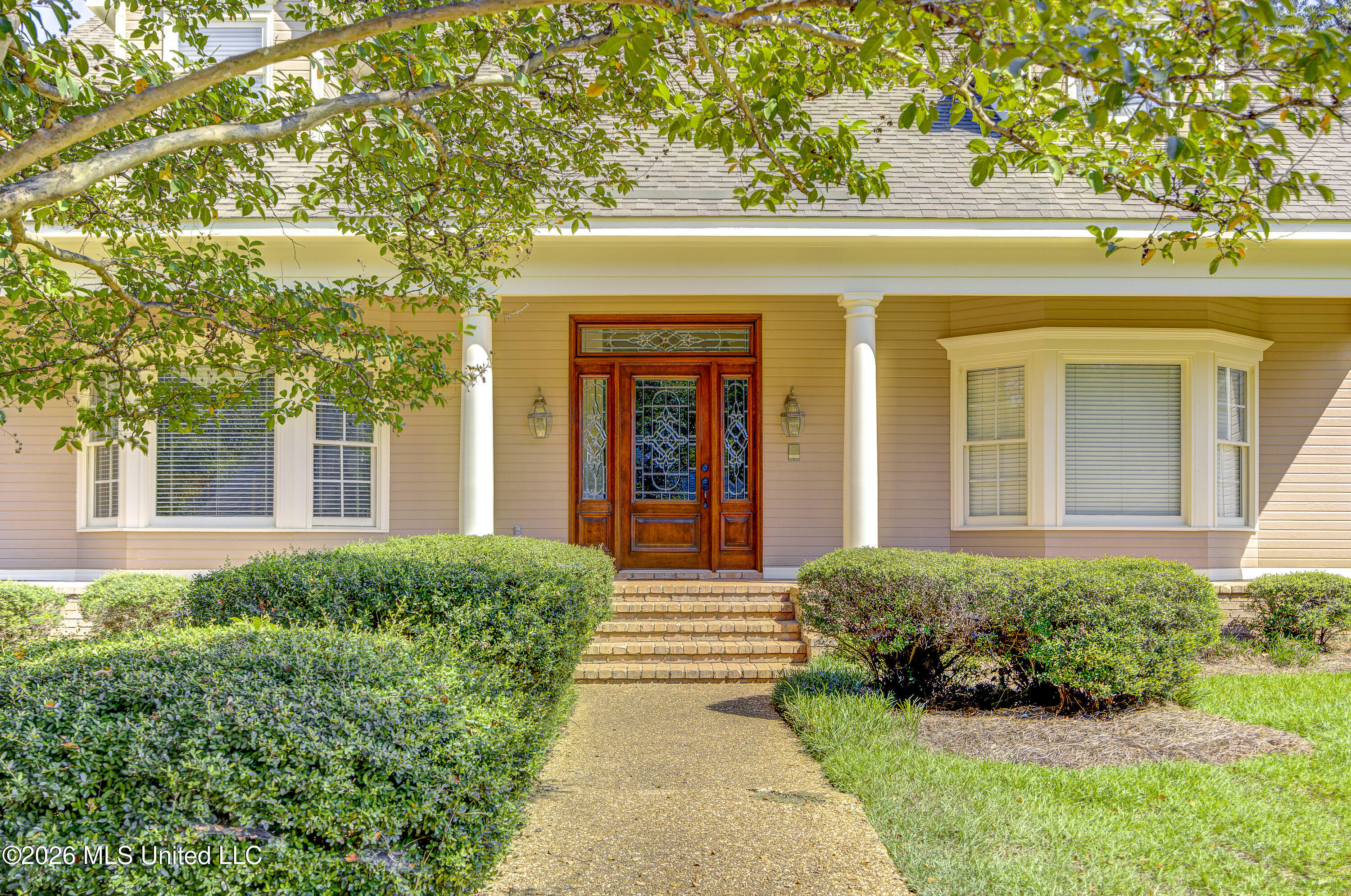 Home entrance with white columns, ornate wood-and-glass door, brick steps, and landscaped shrubs.