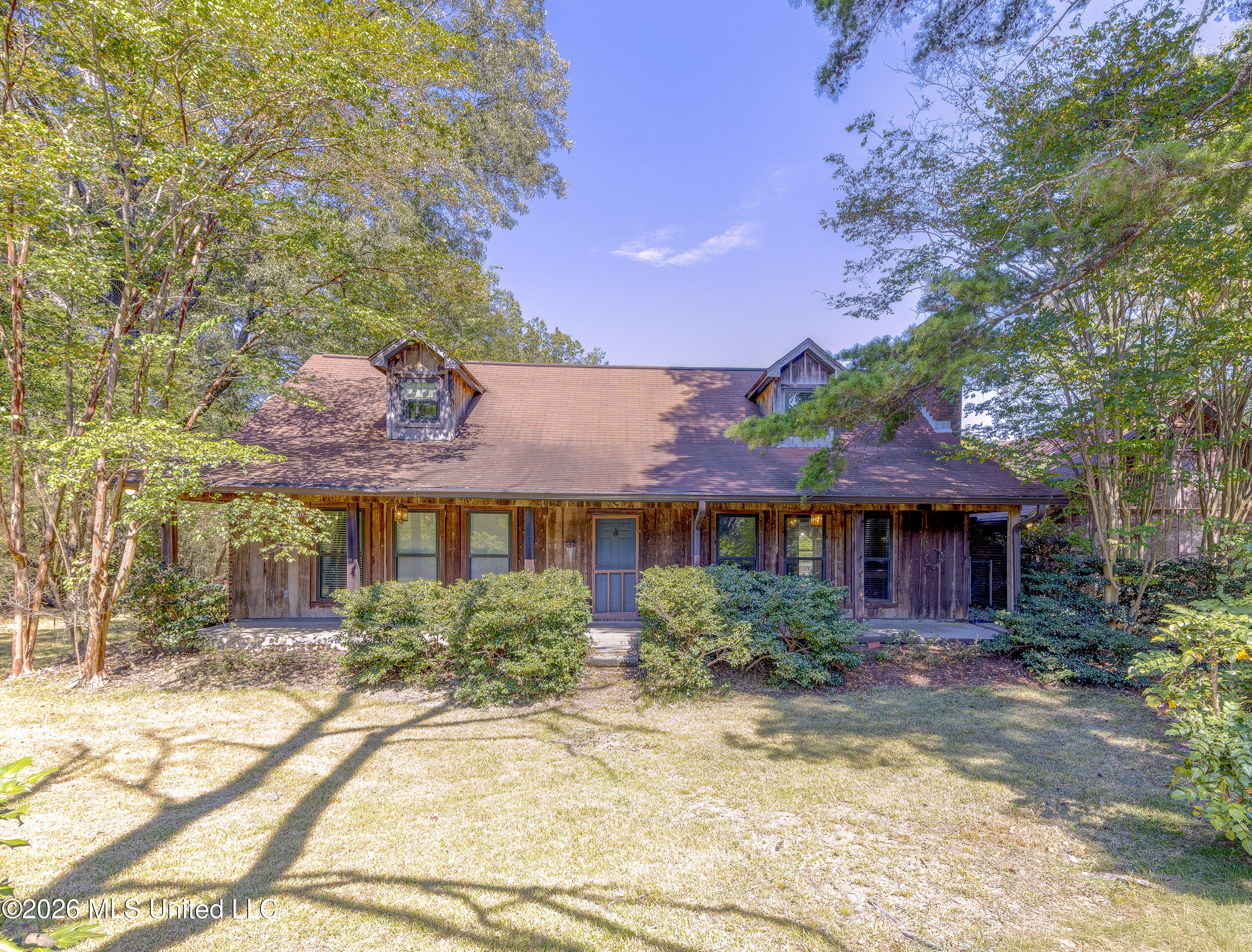Wood-sided house with dormers and a covered porch, surrounded by mature trees and shrubs.