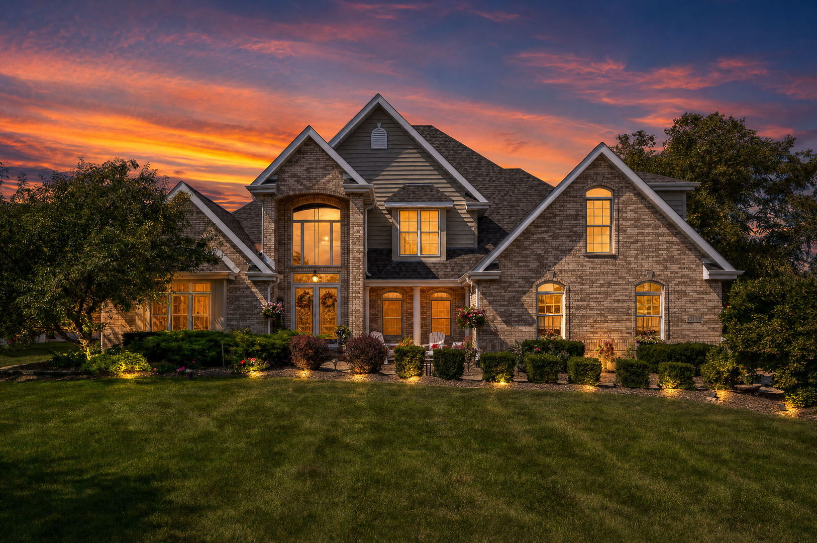 Large brick home with warmly lit windows and landscaped grounds at sunset.