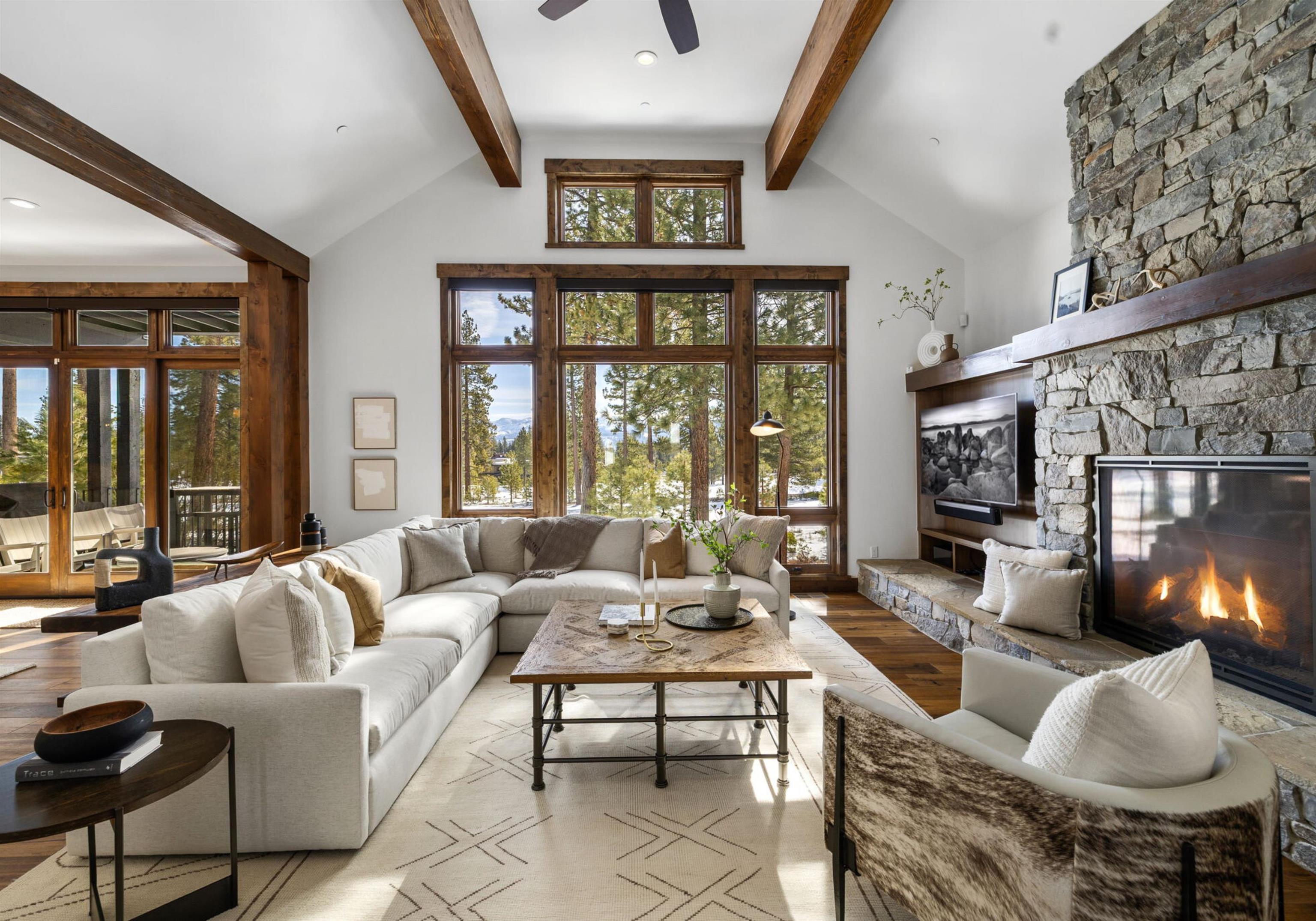 Living room with exposed timber beams, a stone fireplace, and large windows overlooking snowy trees.