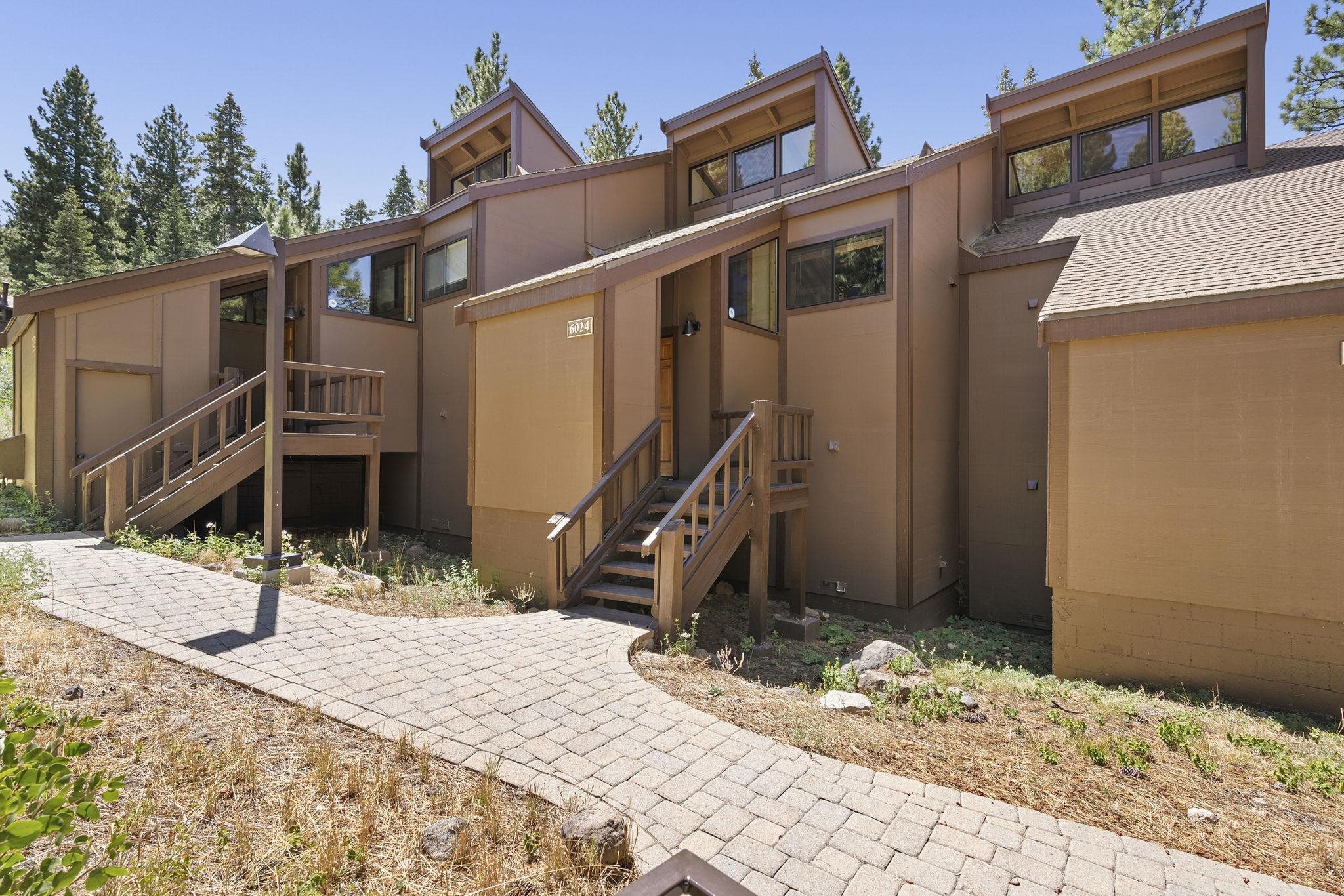 Brown multi-level residence with wooden entry stairs, a paved walkway, and pine trees.