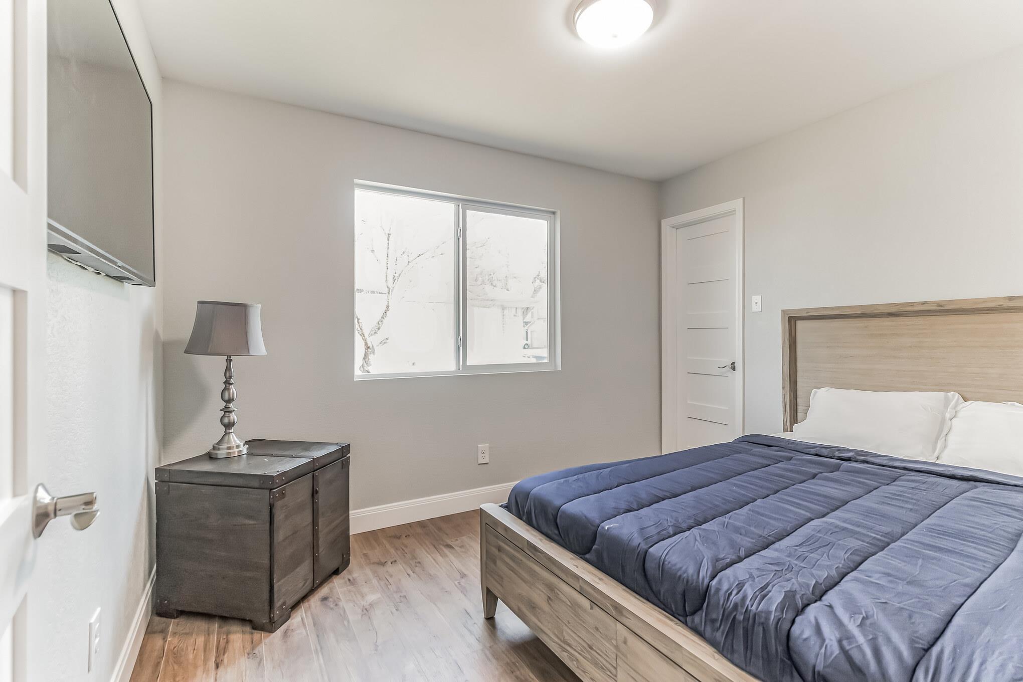 Staged bedroom with a wood bed, blue quilt, dark bedside chest, and window.