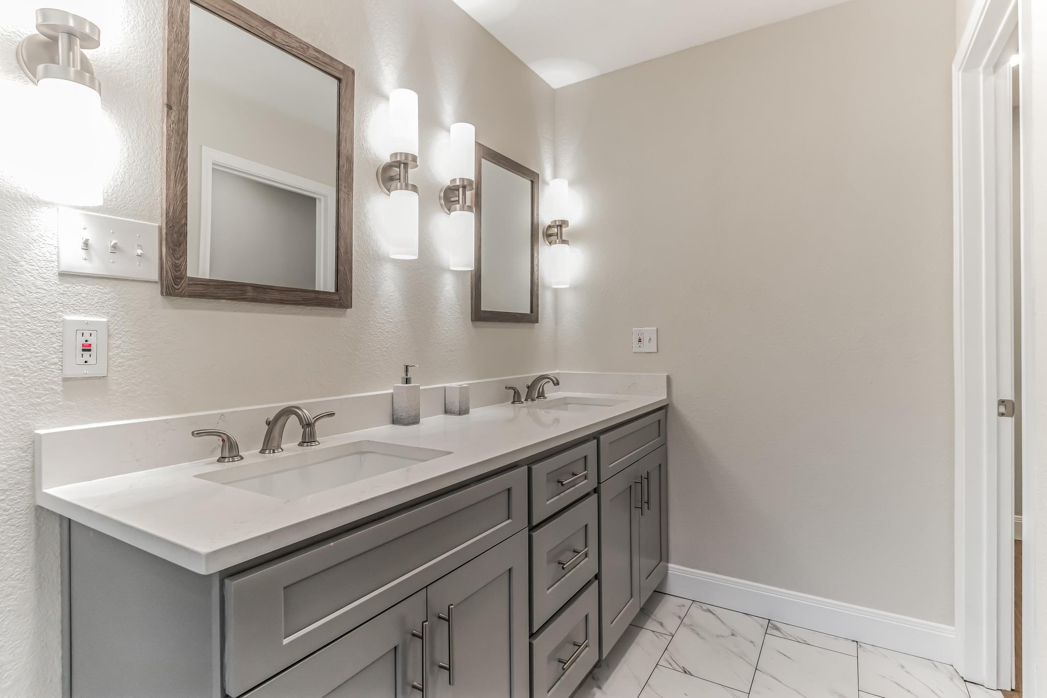 Bathroom with a gray double vanity, white counters, wood-framed mirrors, and marble-look tile floor.