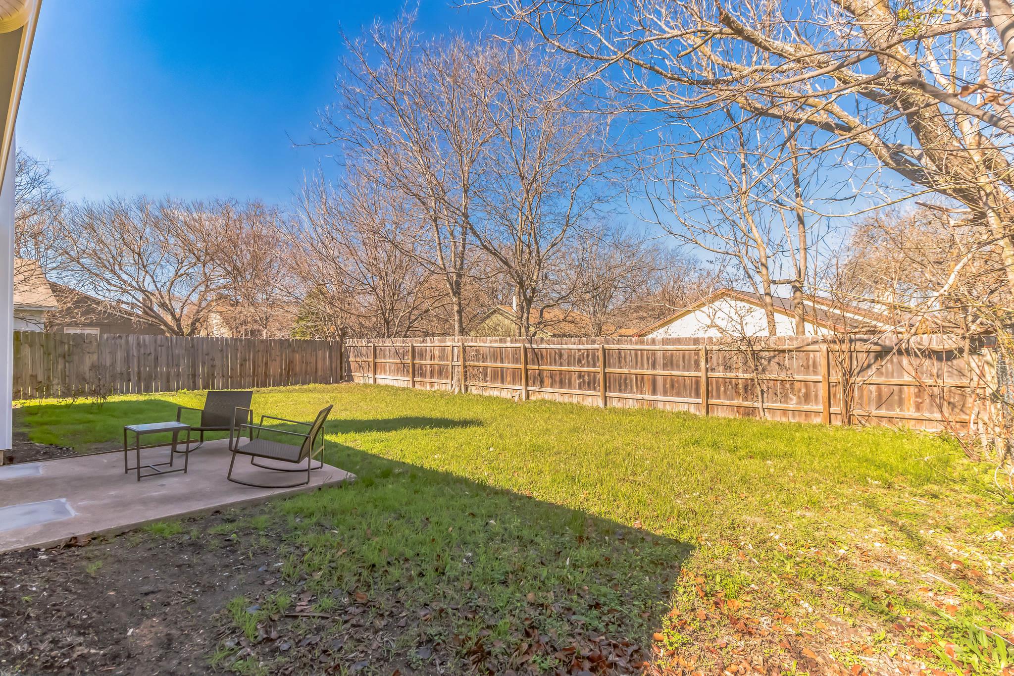 Fenced backyard with a green lawn, patio seating, and leafless trees.