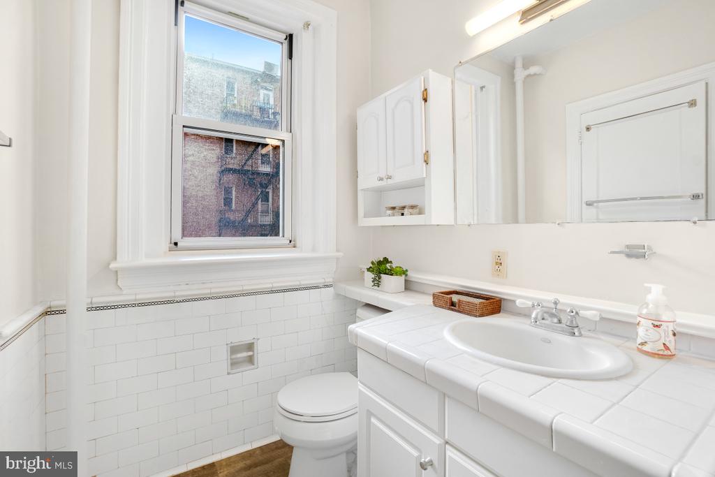 White bathroom with a tiled sink vanity, mirrored medicine cabinet, and window overlooking a brick building.