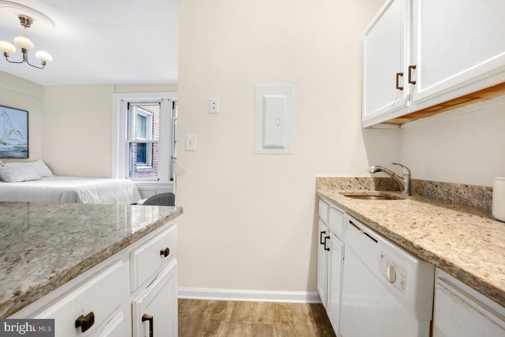 Compact white kitchen with granite counters, a sink, and a view into an adjoining bedroom.