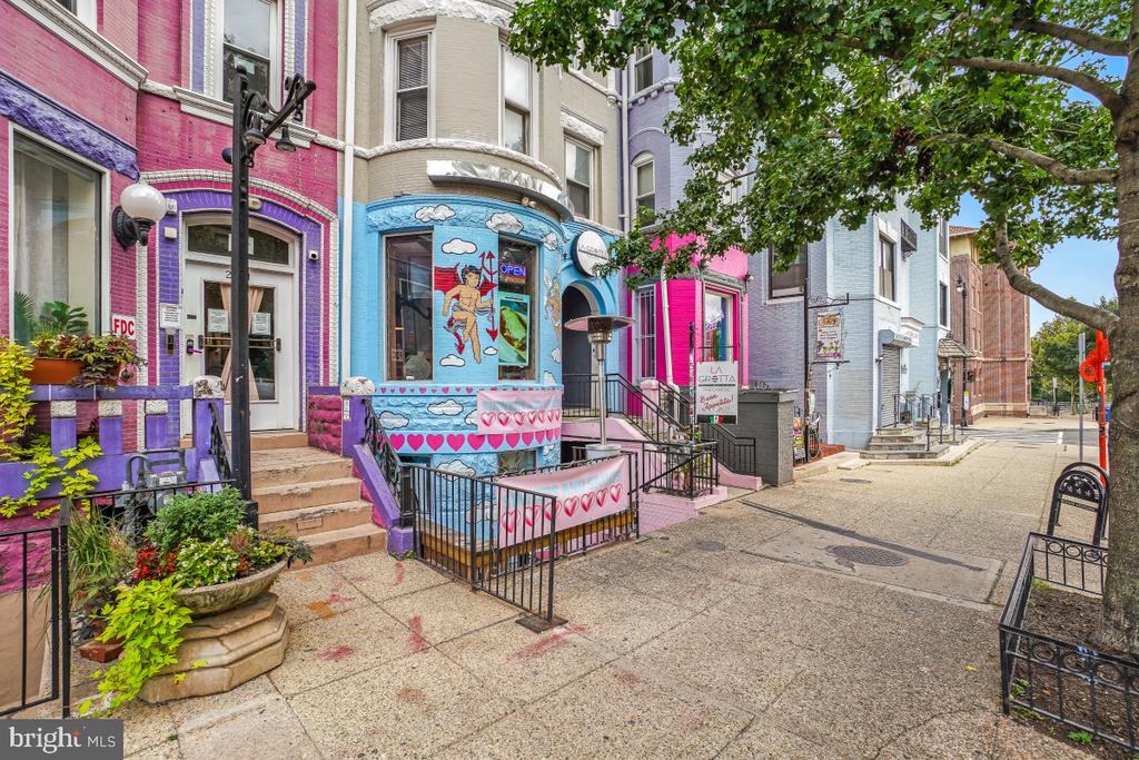 Colorful attached storefronts with turquoise and pink facades line a tree-shaded sidewalk.