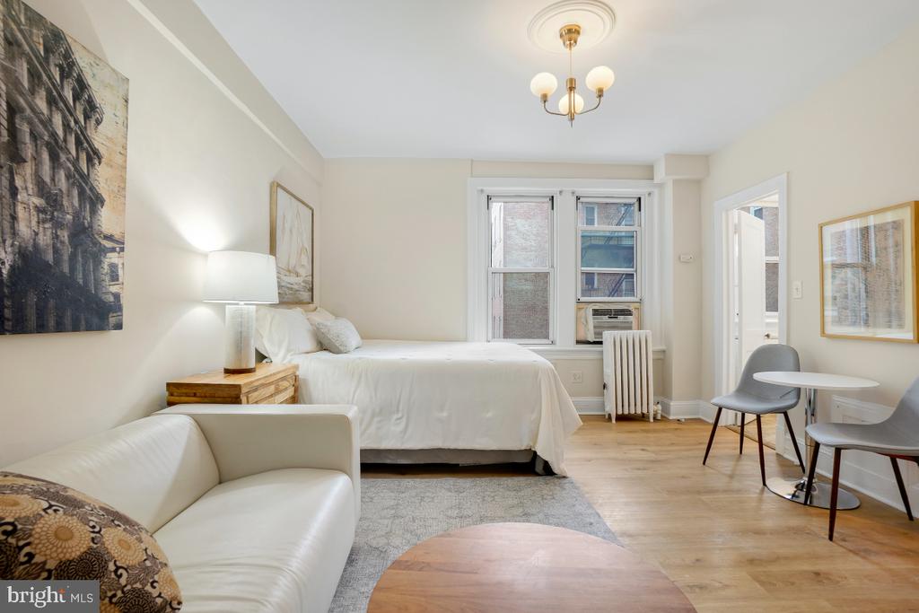 Furnished studio bedroom with a white bed, sofa, and dining nook beside tall windows.