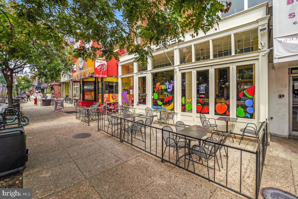 Storefronts and sidewalk café seating beneath leafy trees on a sunny city street.