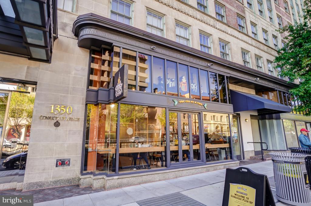 Stone building exterior with a glass-fronted café, sidewalk seating, and a curbside-pick-up sign.