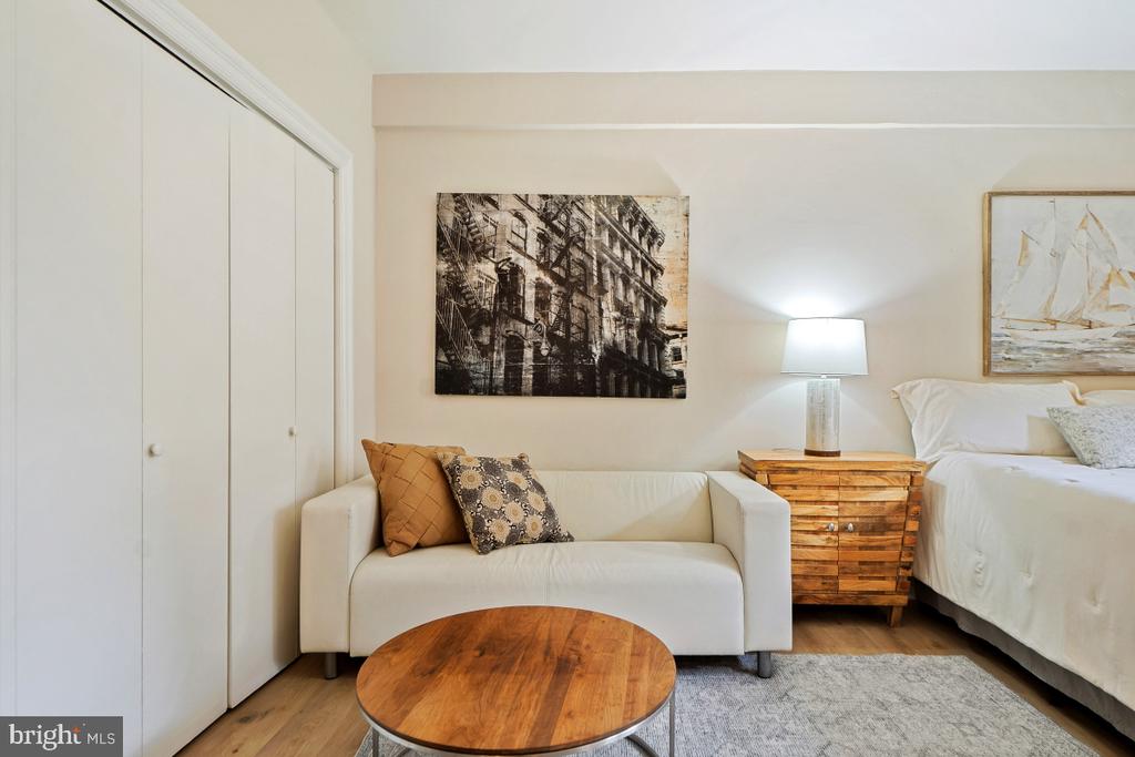 Furnished studio bedroom with a white bed, compact sofa, and round wooden coffee table.