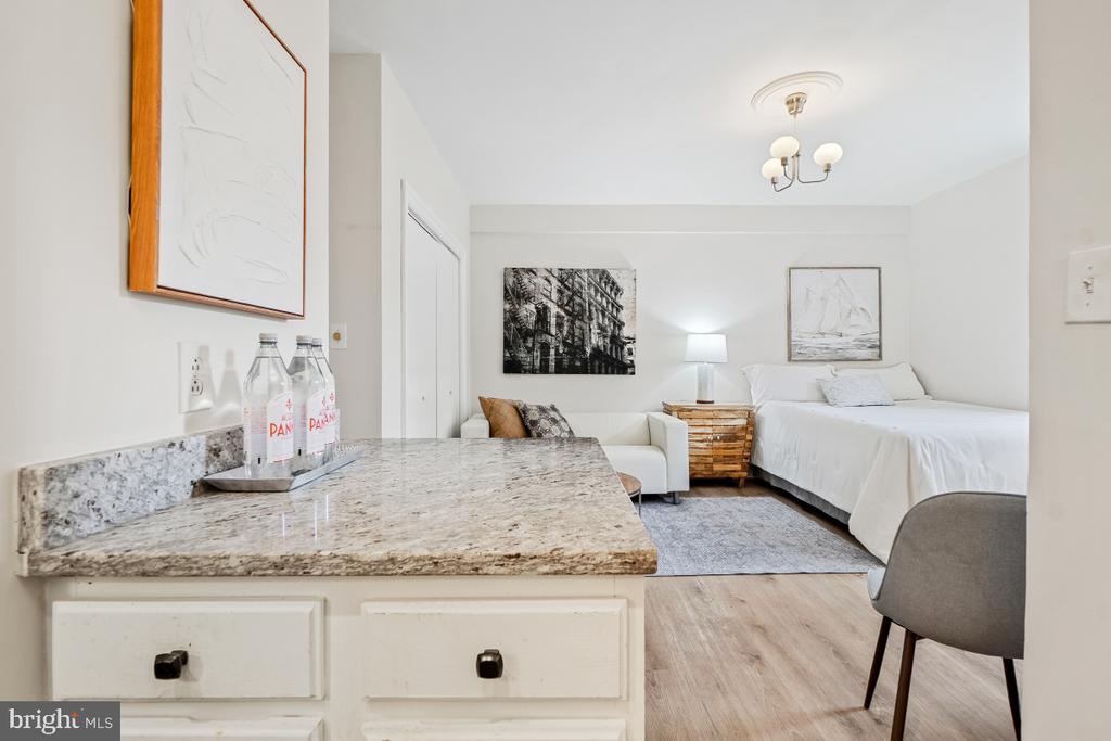 Furnished studio bedroom with a white bed, small sofa, and granite-topped cabinet in the foreground.