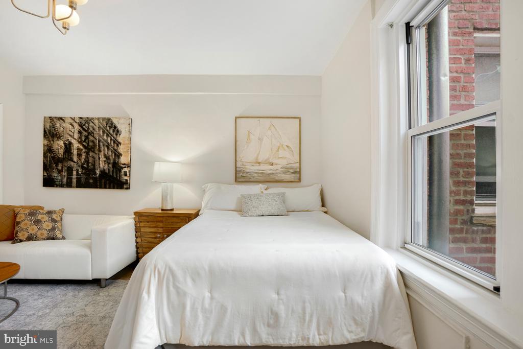 Bedroom with a white bed, sofa, and tall windows beside a brick wall.