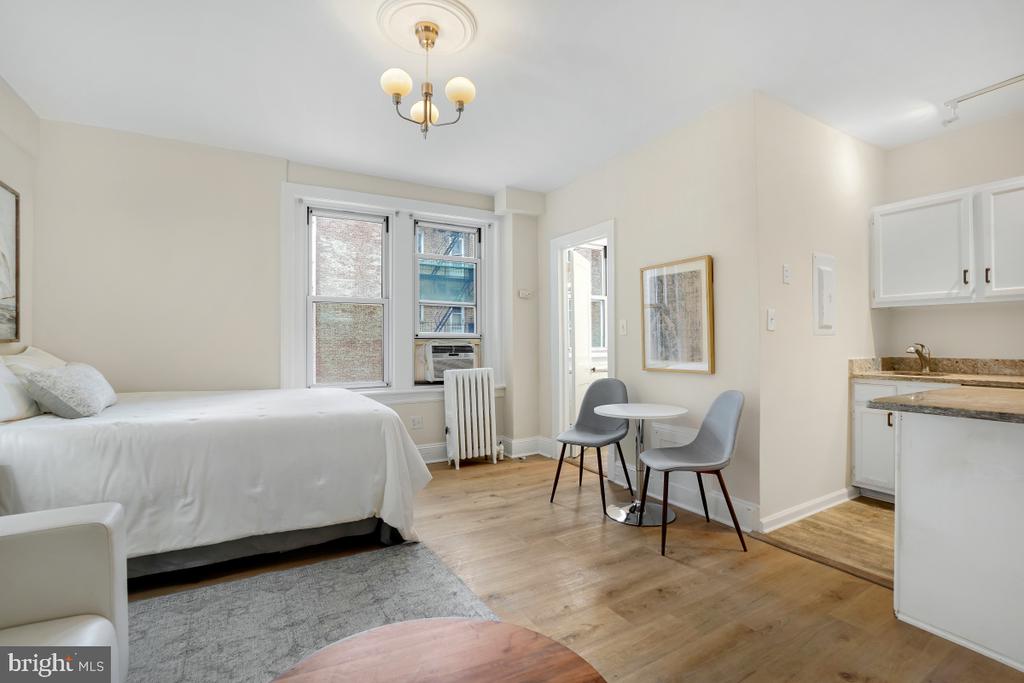 Studio apartment with a bed, compact kitchenette, and seating area beside large windows.
