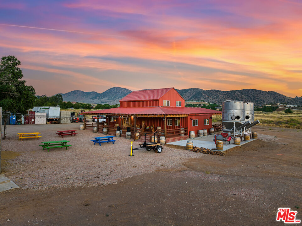Red barn-style building with picnic tables and metal silos beneath a colorful sunset sky.