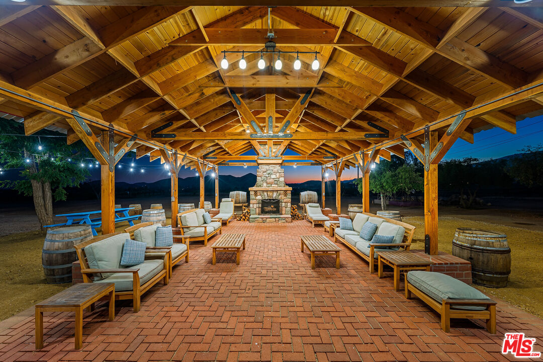 Covered outdoor patio with timber pavilion, brick flooring, cushioned seating, and a stone fireplace.
