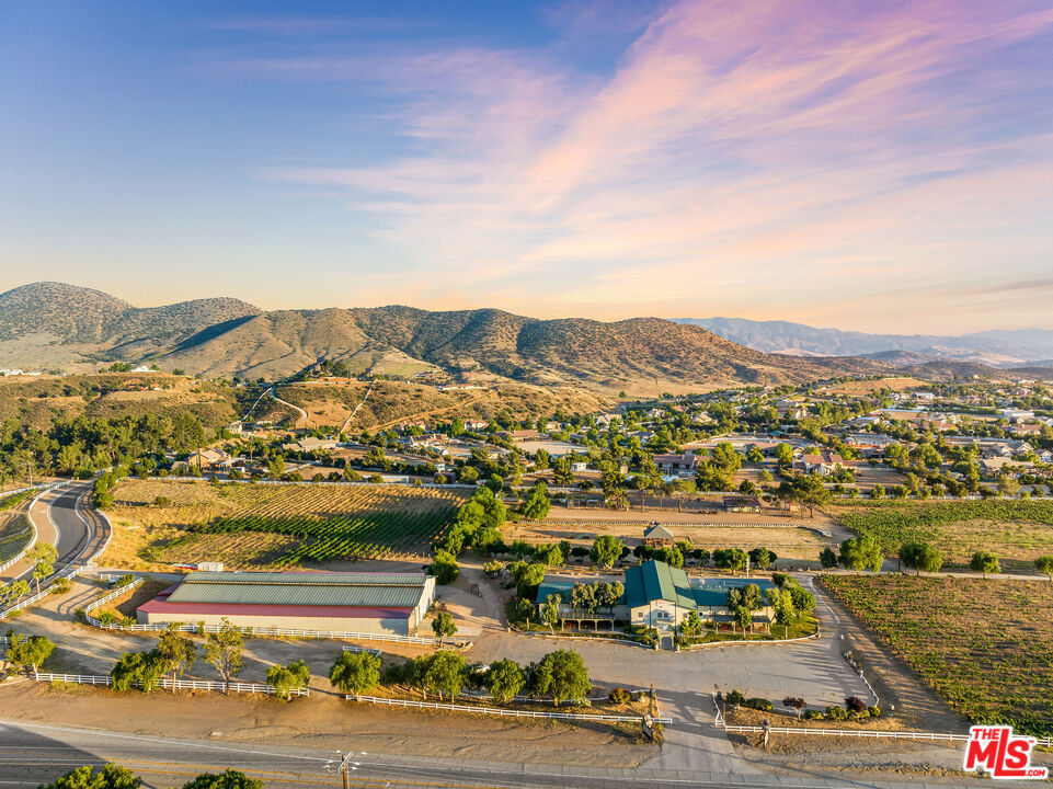 Aerial view of a ranch property with barns, fenced grounds, and mountain foothills at sunset.