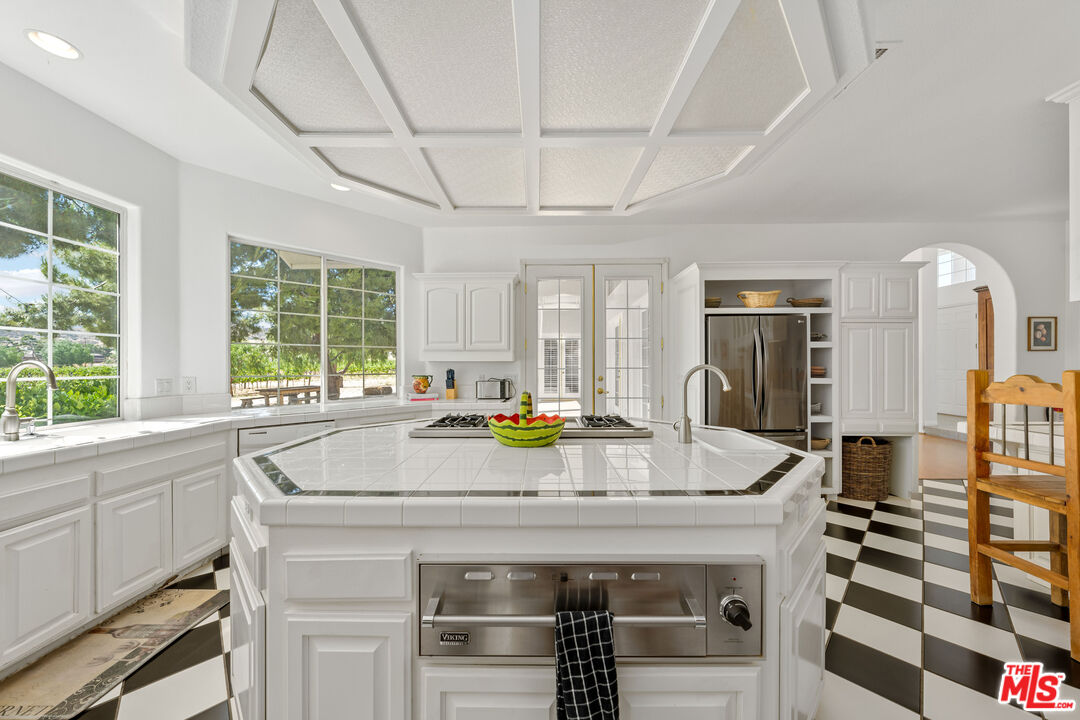 Bright white kitchen with a large tiled island, coffered ceiling, and black-and-white checkered floor.