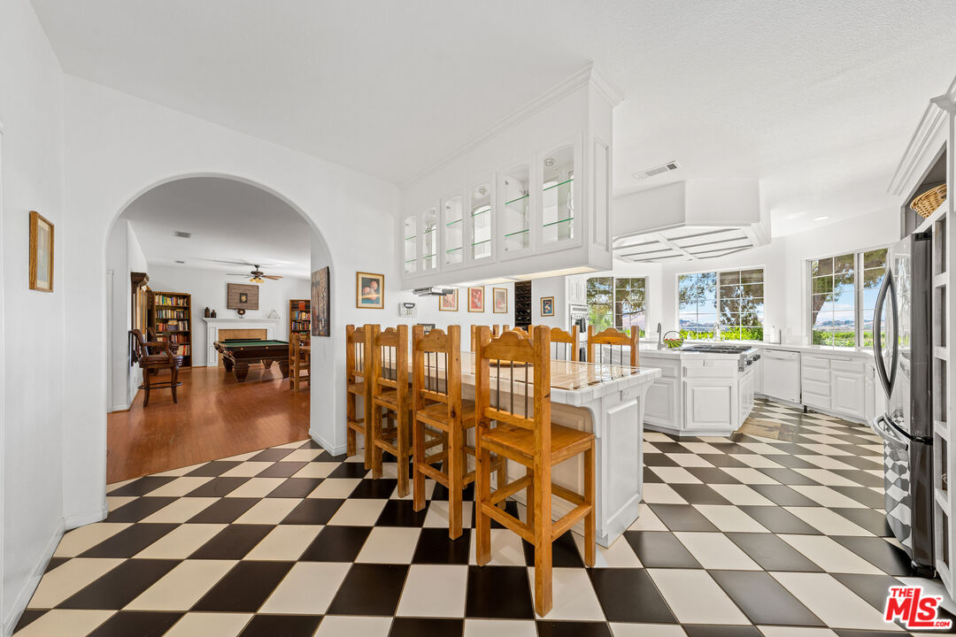 Bright white kitchen with checkerboard floors and wood stools, opening to a billiards room.
