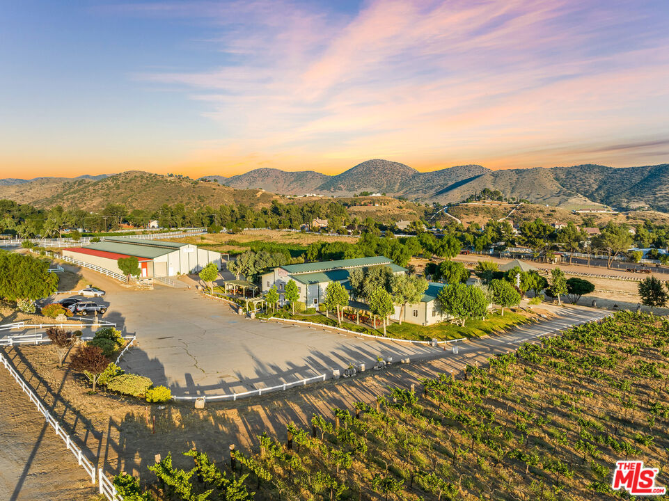 Aerial view of a rural property with green-roofed buildings, vineyard rows, and surrounding mountains.