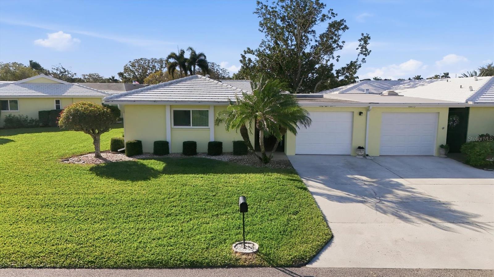 Pale yellow single-story home with two garage doors, a broad lawn, and palm trees.