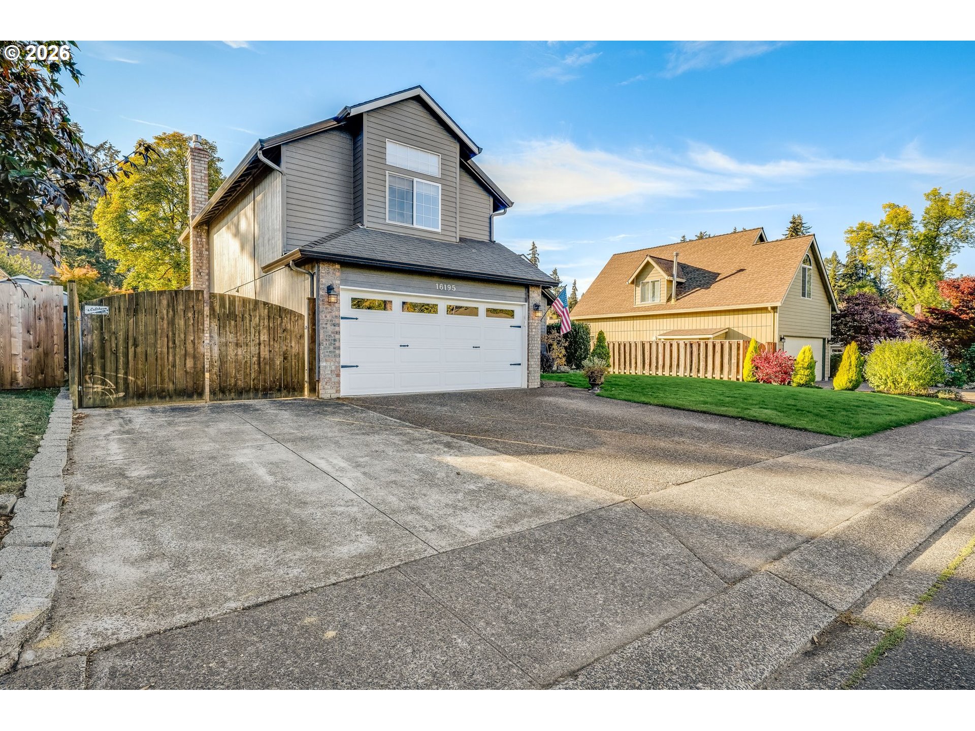 Two-story gray house with a white garage door, broad driveway, and wooden fencing.