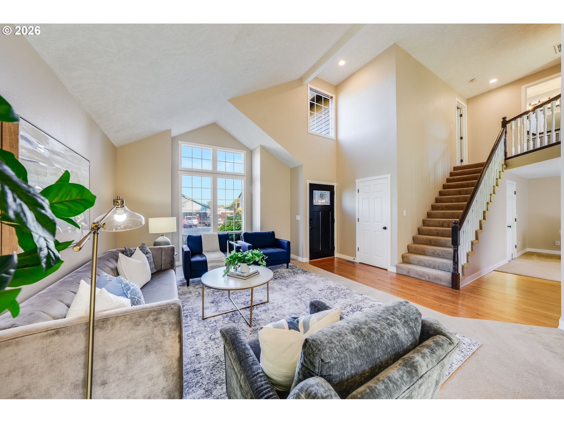 Staged living room with vaulted ceilings, tall windows, and a staircase beside the seating area.