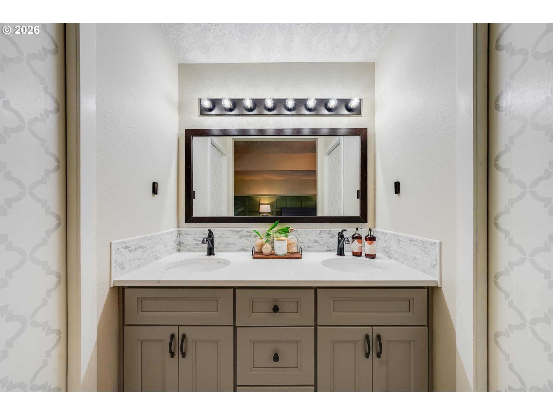 Bathroom vanity with two sinks, taupe cabinetry, tiled backsplash, and a large framed mirror.