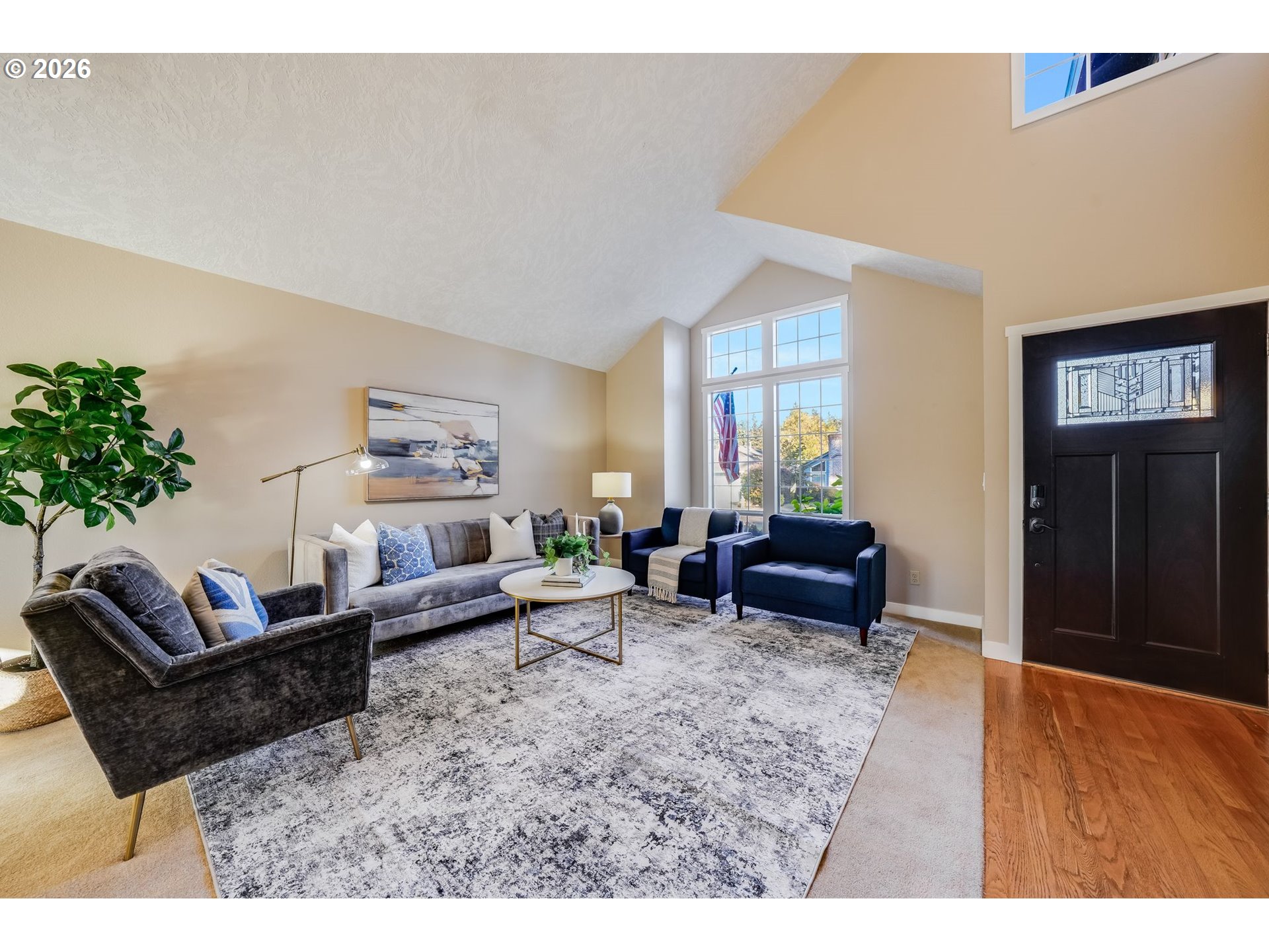 Furnished living room with vaulted ceiling, large windows, and a dark front door.