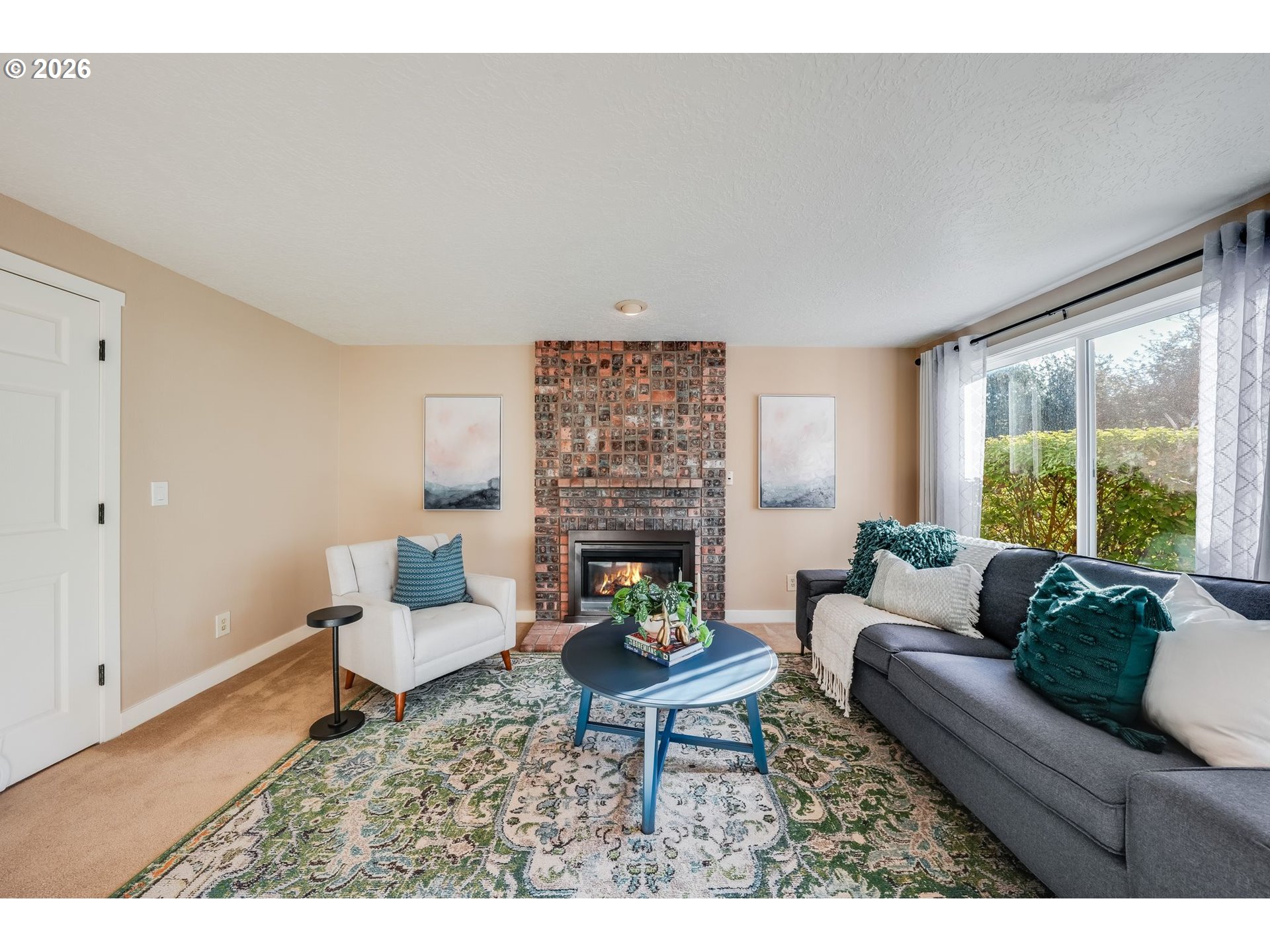 Living room with a brick fireplace, gray sofa, and large windows overlooking greenery.
