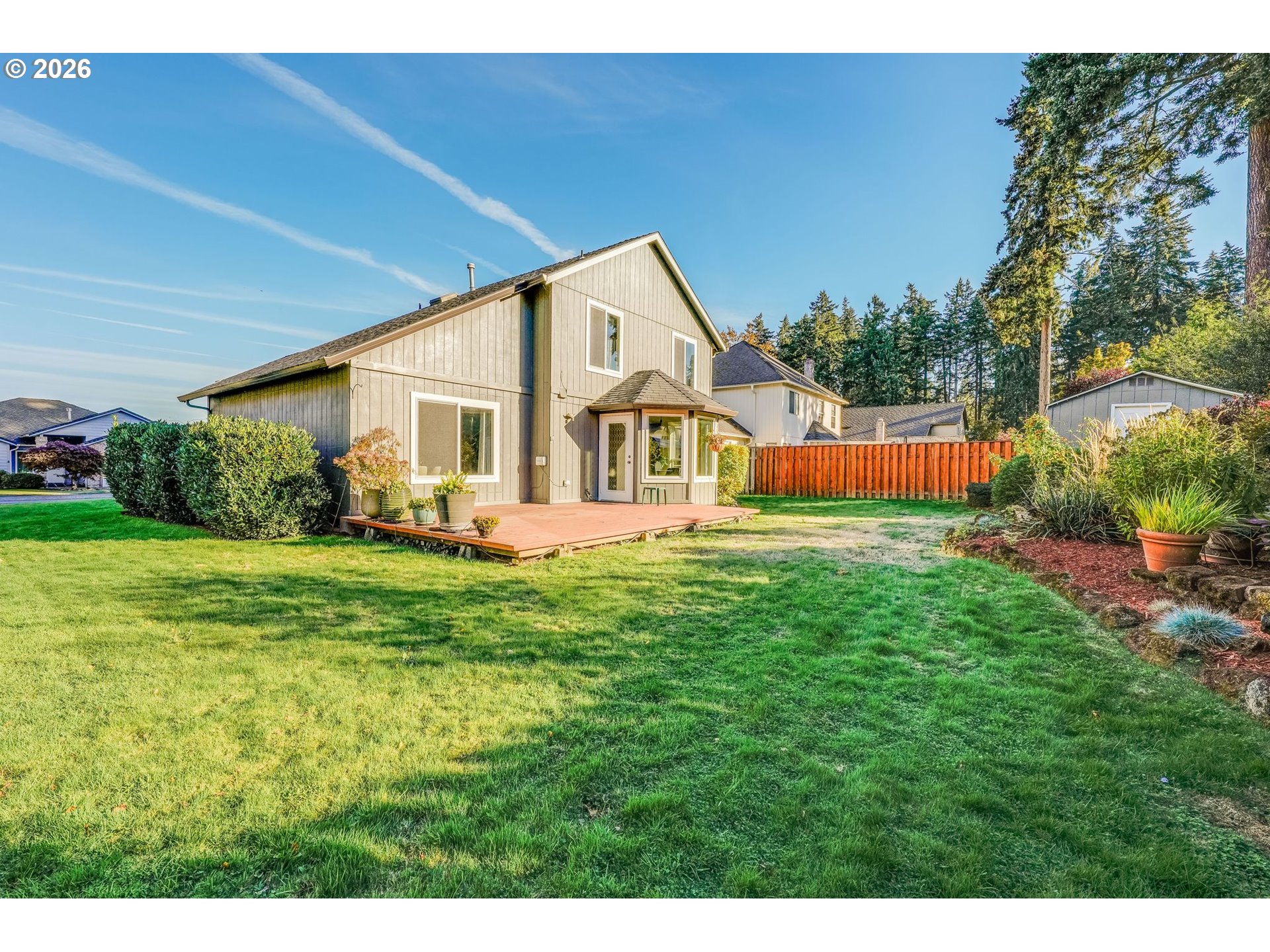 Rear of a two-story beige home with a wooden deck overlooking a broad lawn and red fence.