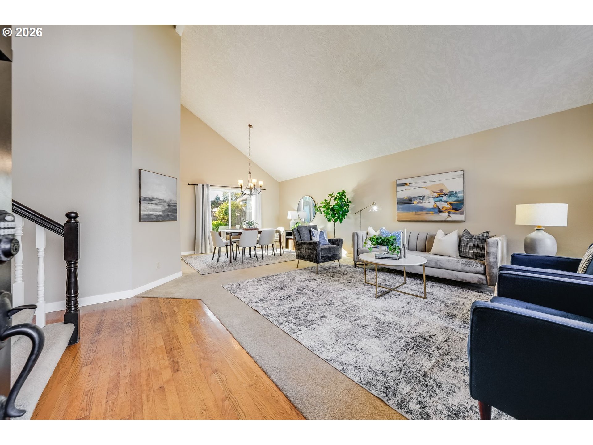 Staged living and dining room with a vaulted ceiling, gray seating, hardwood entry, and bright window.