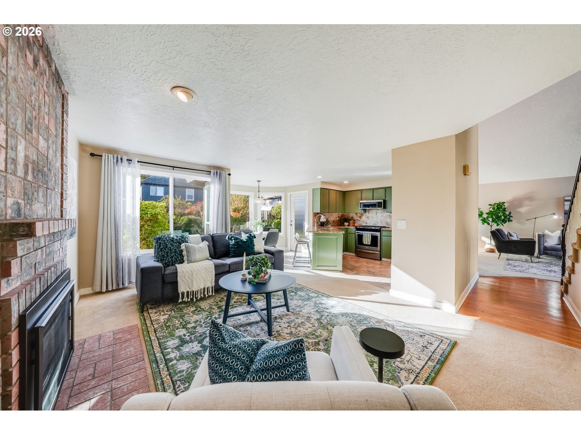 Open-plan living room with brick fireplace, patterned rug, and green kitchen cabinets.