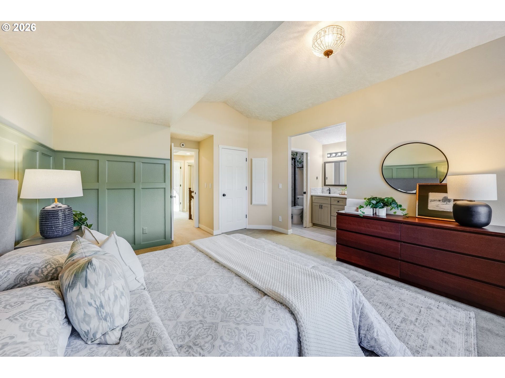 Staged bedroom with green paneled wainscoting, patterned bedding, and an oval mirror above a dark wood dresser.