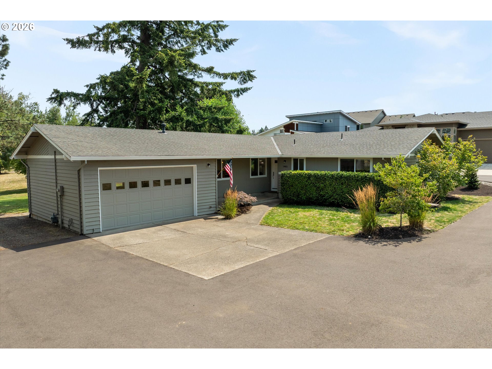Single-story gray home with an attached two-car garage, front hedge, and American flag.