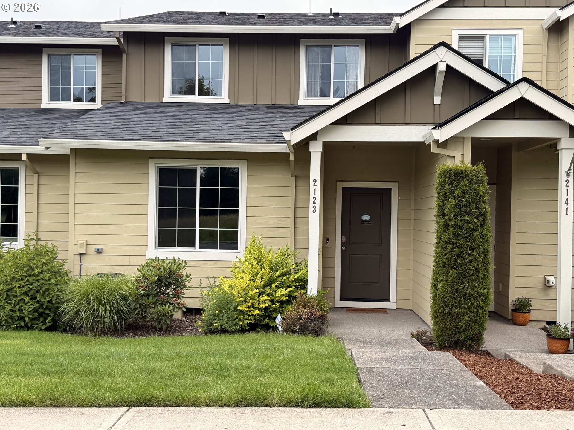 Beige two-story attached homes with covered entries, landscaped shrubs, and a concrete walkway.