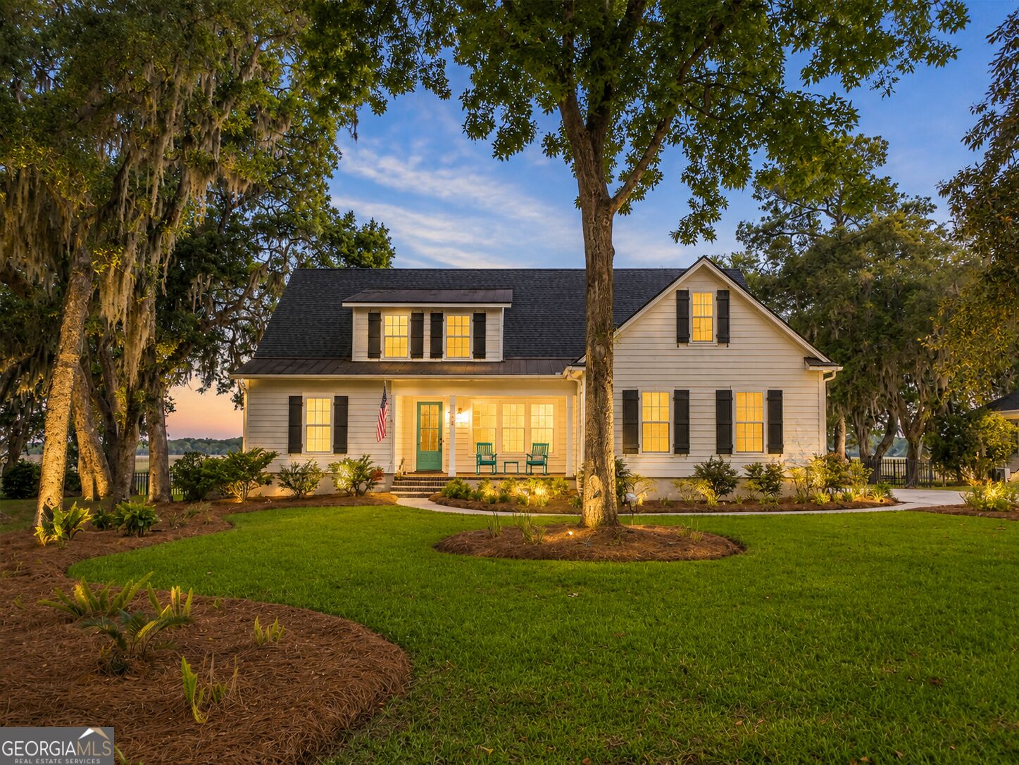 Two-story home with black shutters and a green door, framed by mature trees at dusk.