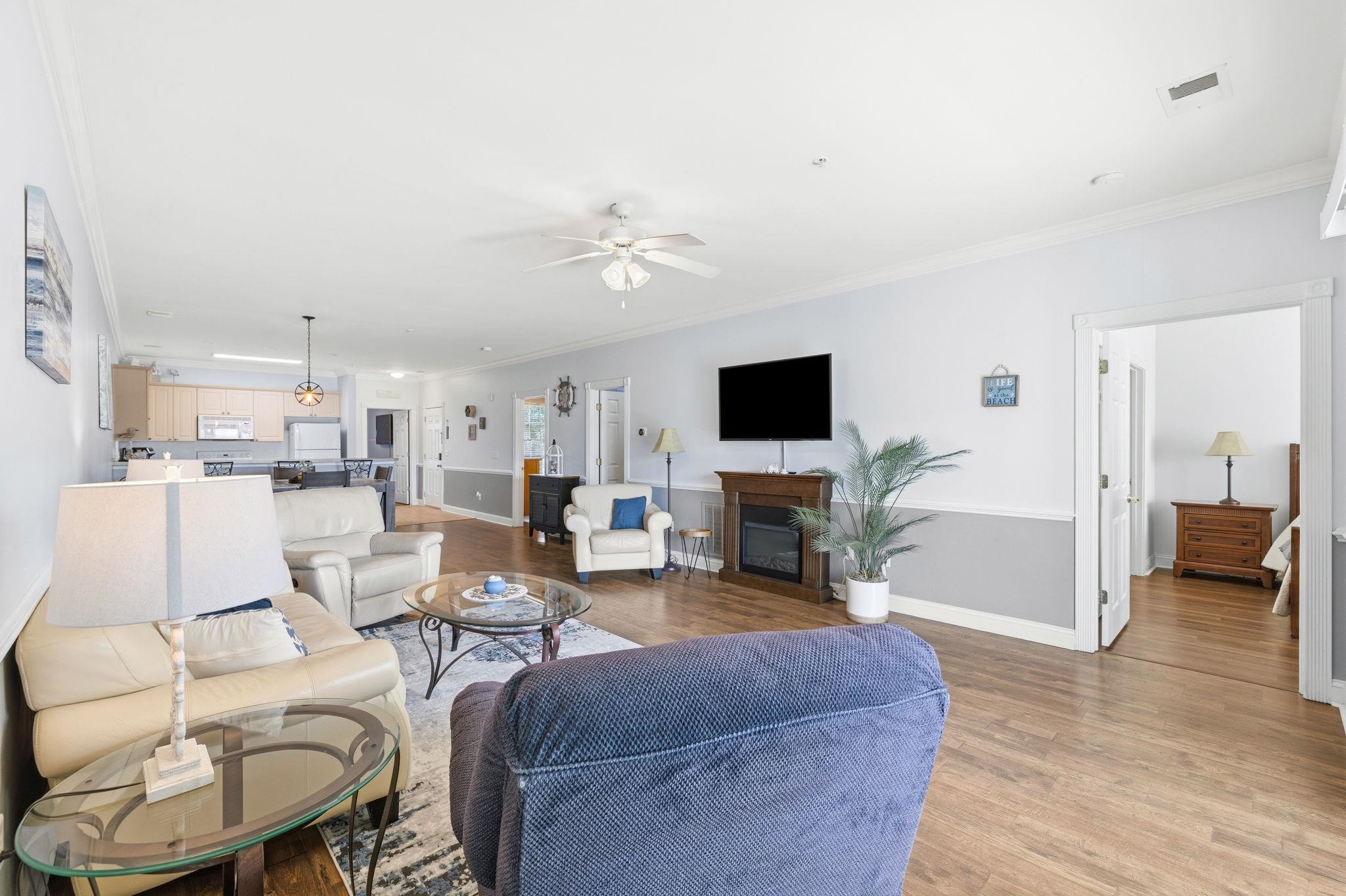 Furnished living room opening to a kitchen, with cream leather seating and a fireplace beneath a wall-mounted TV.