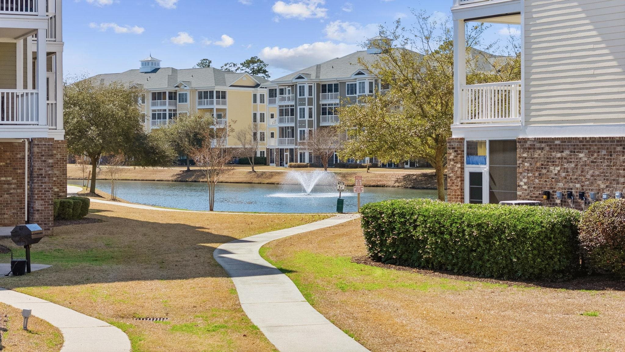 Residential buildings across a pond with a fountain and curving sidewalk in the foreground.