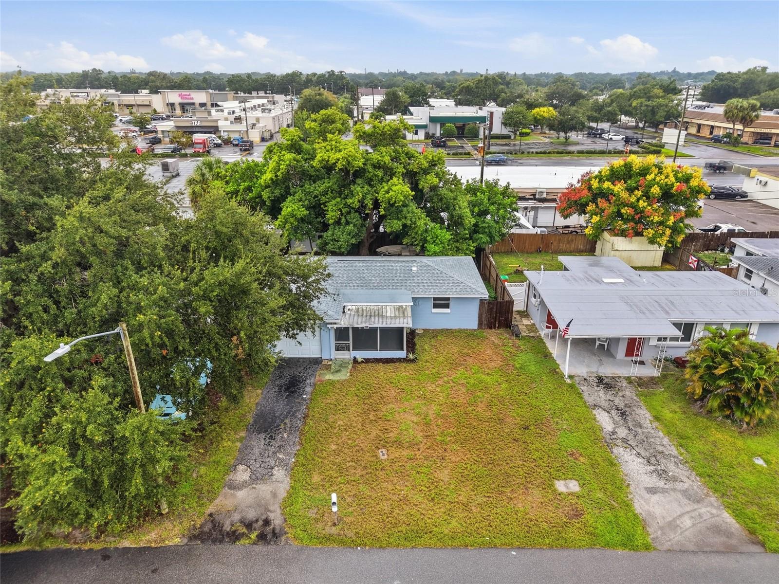 Aerial view of a blue house with a screened porch and overgrown front yard beside commercial buildings.