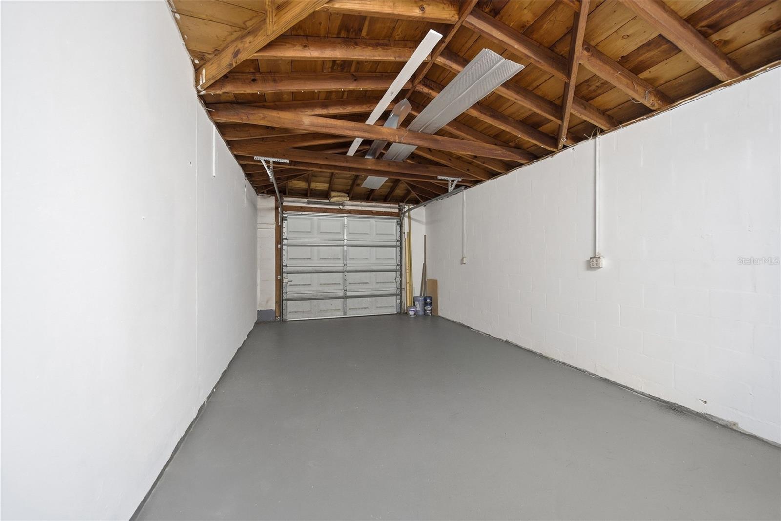 Empty garage with exposed wooden rafters, white walls, and a gray concrete floor.