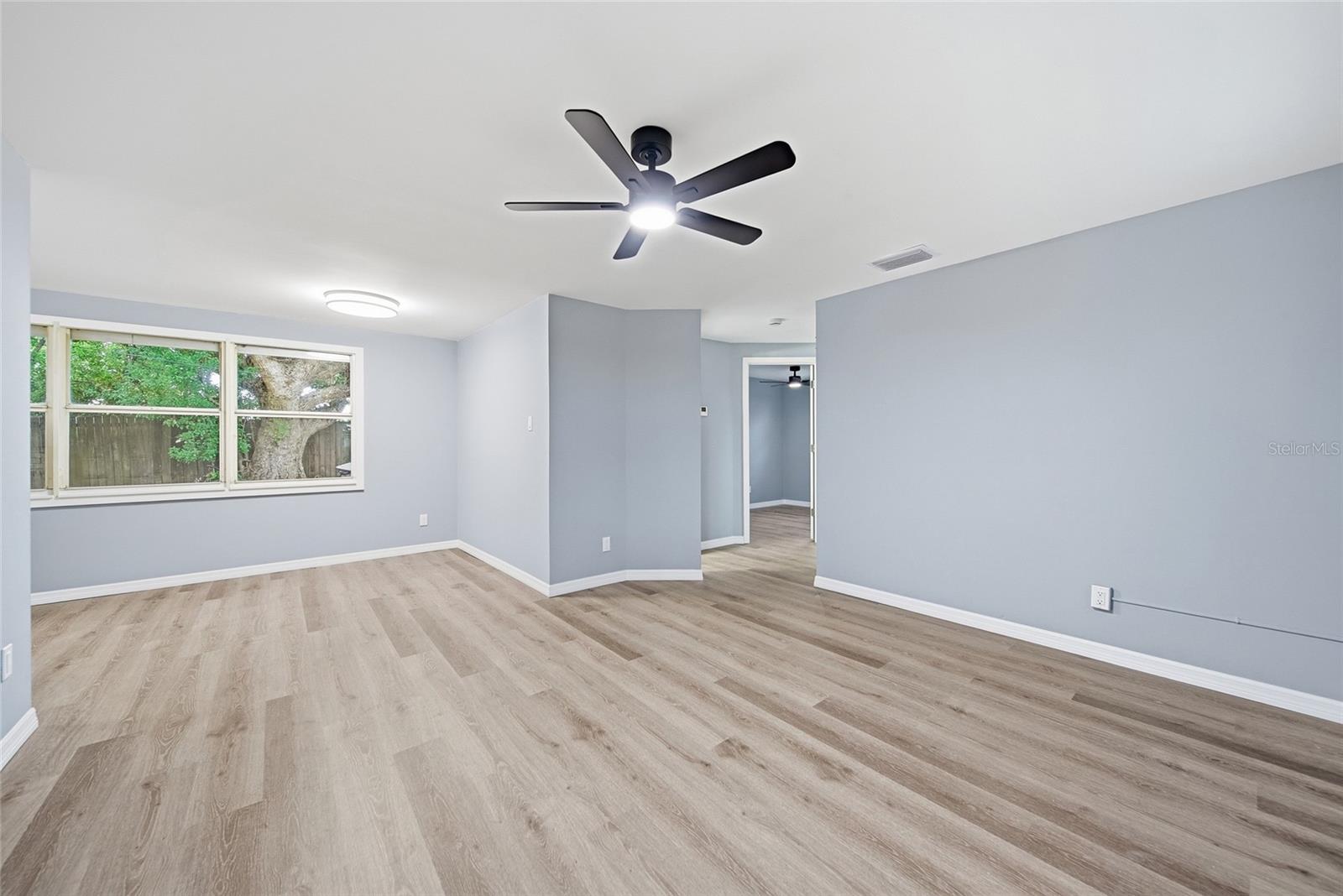 Vacant living room with wood-look flooring, a black ceiling fan, and wide windows overlooking trees.