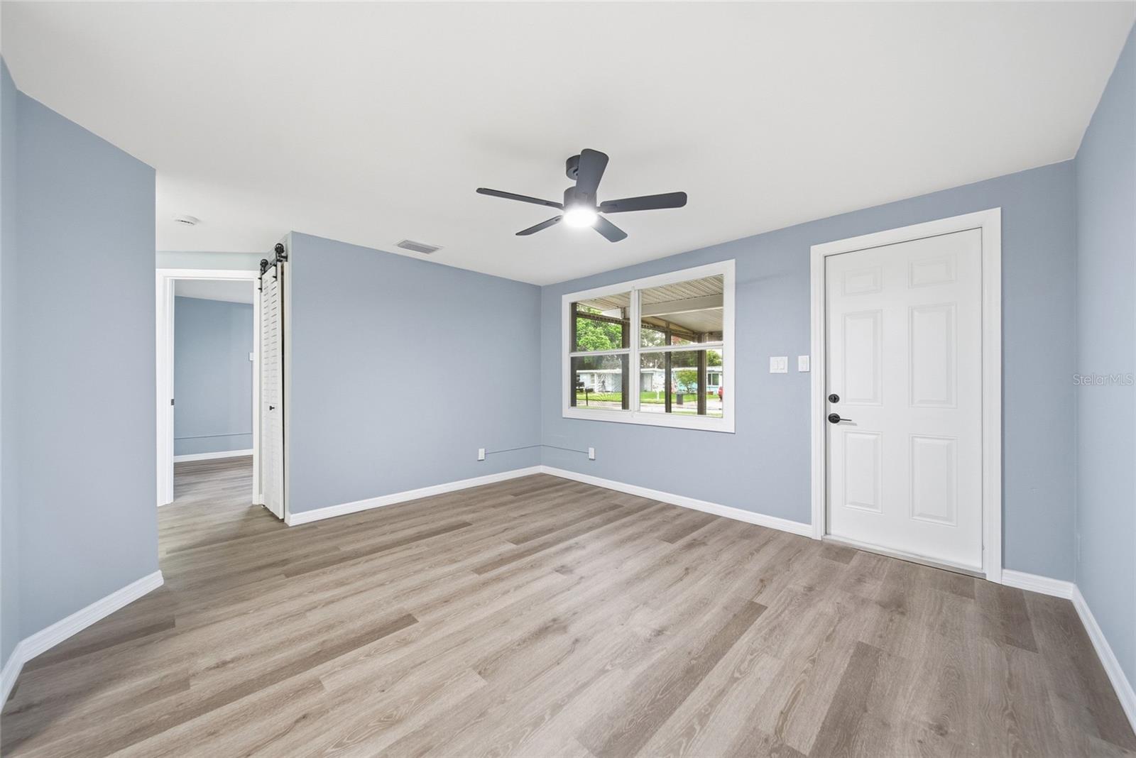 Empty room with pale blue walls, gray plank flooring, a black ceiling fan, and a window.