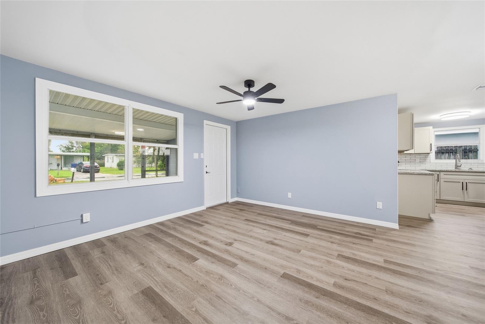 Empty living room with pale blue walls, gray wood-look floors, large windows, and an adjoining kitchen.