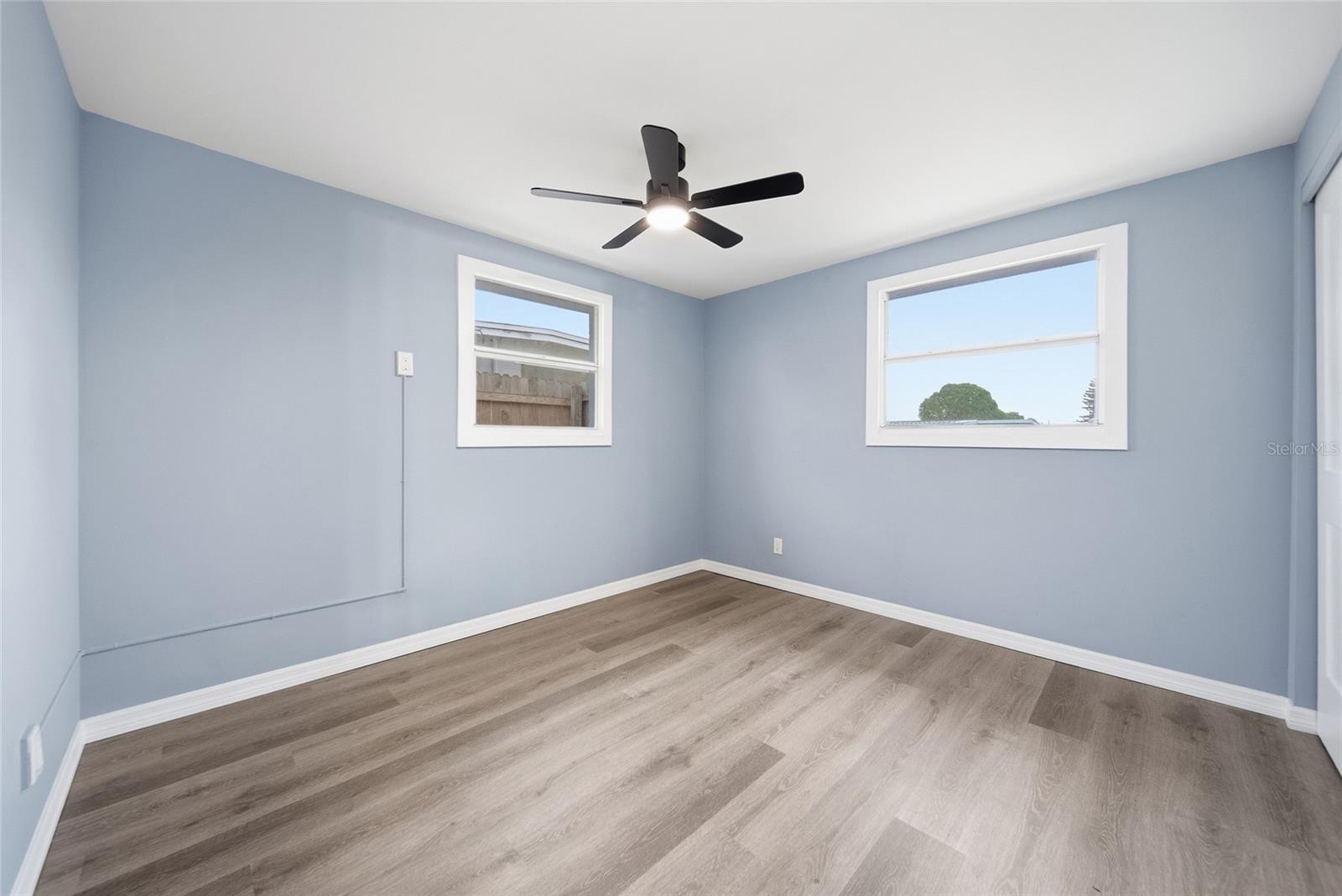 Vacant room with blue walls, wood-look flooring, two windows, and a black ceiling fan.