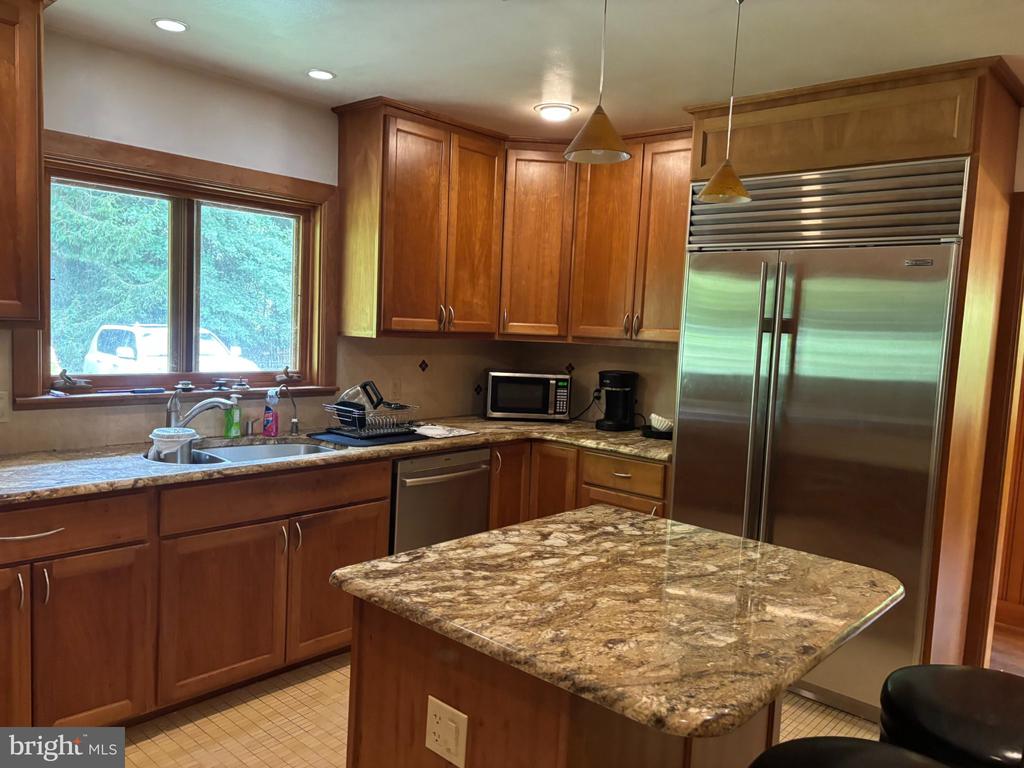 Kitchen with wood cabinetry, granite island, stainless steel refrigerator, and a wide window above the sink.