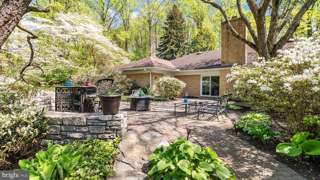 Backyard patio with outdoor furniture, a stone retaining wall, and flowering trees beside a brick house.