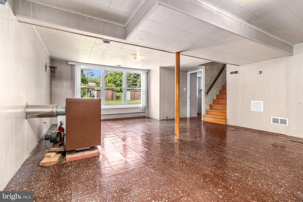 Empty lower-level room with a large yard-facing window, wood support post, and staircase.